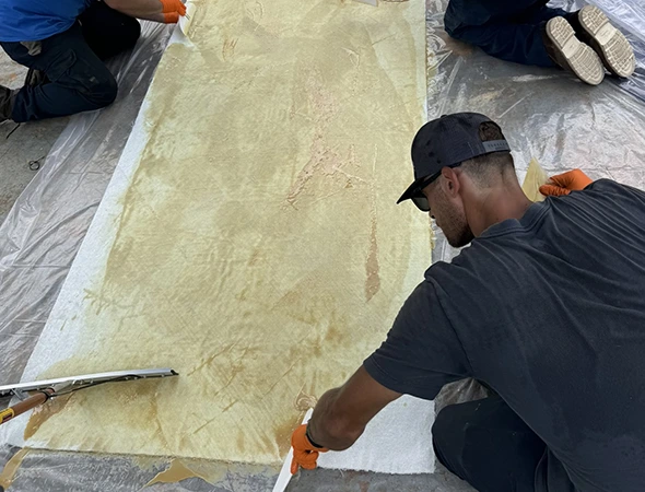 Three people work on a large, beige canvas, smoothing it with tools. One wears a cap and gloves. The setting appears collaborative and focused.