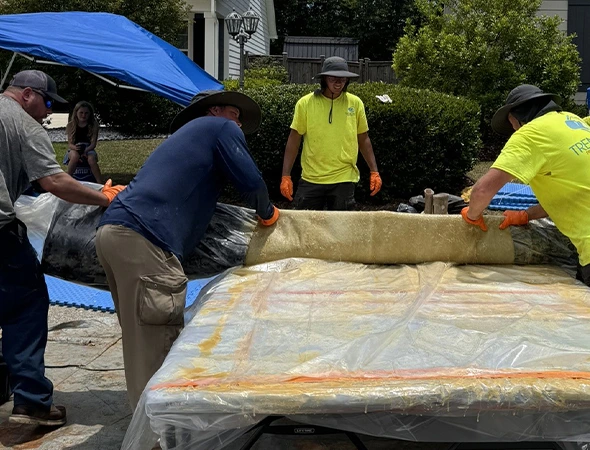 Four workers in hats and gloves unroll fibreglass insulation on a table outdoors. A blue tarp and greenery are visible, creating a sense of teamwork.