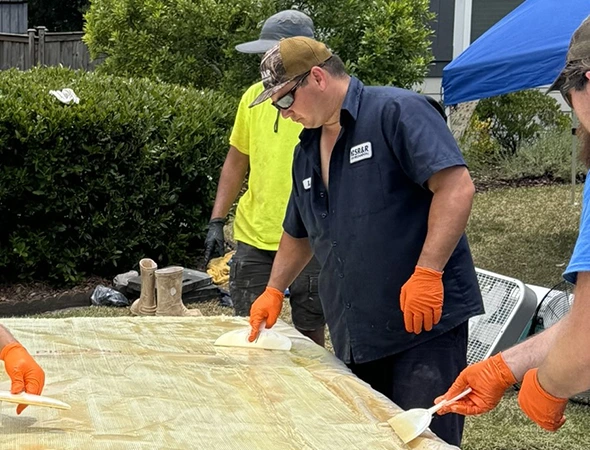 Three people work outdoors, applying resin to a flat surface. Wearing gloves and casual outfits, they focus intently. Greenery and a canopy surround them.