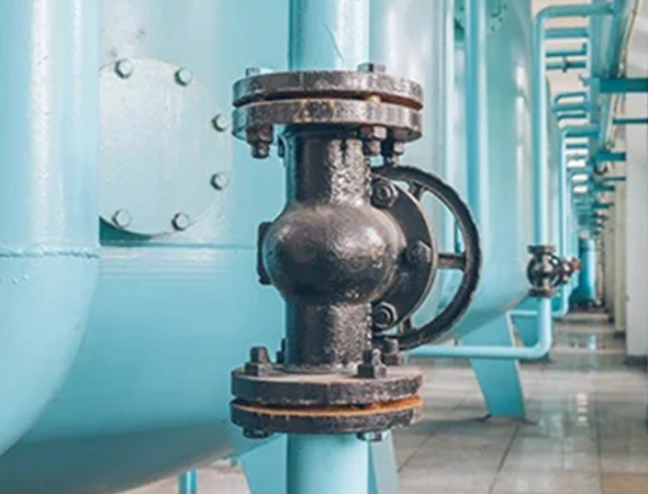 Close-up of a large industrial black valve on a light blue pipe in a factory setting. The floor is reflective, creating a clean, orderly atmosphere.