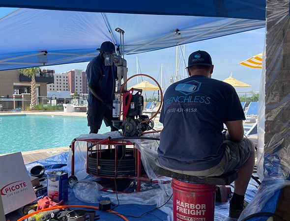 Two workers under a canopy perform maintenance near a pool. One operates machinery, the other sits on a tool bucket. Background features a sunny poolside area.