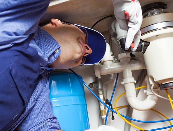 A plumber in a blue shirt and cap works under a sink, using tools in a confined space. He appears focused, surrounded by pipes and a blue tank.