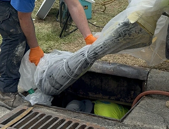 Two workers in gloves handle a large wrapped object being lowered into a storm drain. One worker in a neon shirt is partially visible below.