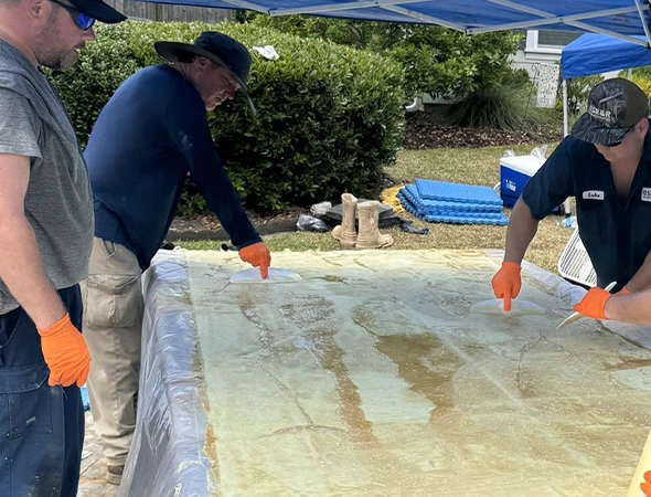 Three people work under a canopy, layering fiberglass on a large surface. They wear gloves and focus intently, conveying a collaborative and industrious mood.