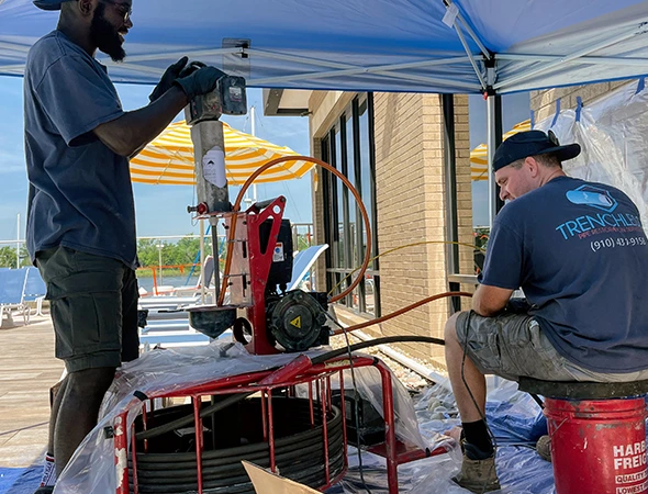 Two workers under a blue canopy operate a red machinery setup. One stands, focusing on the equipment, while the other sits on a bucket, appearing attentive.