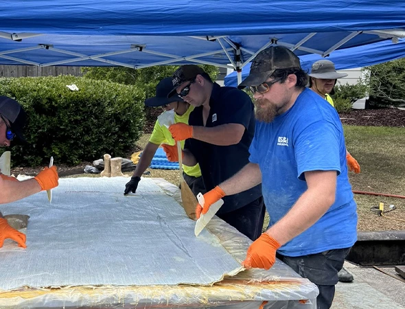 Four men in blue shirts and orange gloves collaborate on a large plastic sheet, demonstrating trenchless plumbing techniques.