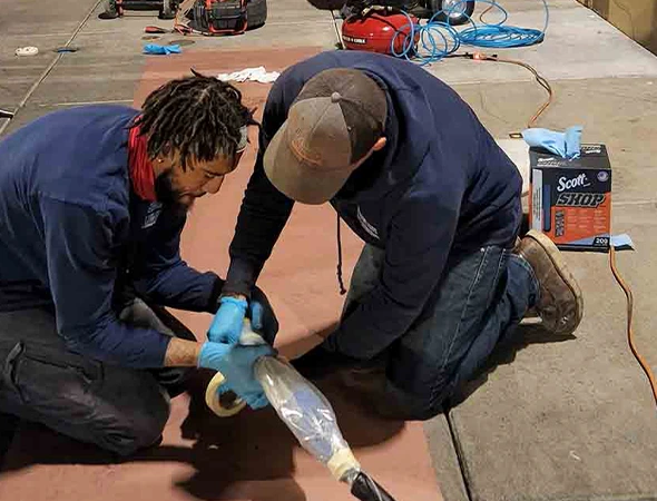 Two men wearing gloves and work clothes are kneeling on the floor, engaged in a focused task. They appear to be wrapping or repairing an object with clear plastic and tape. A box of shop towels and tools are visible in the background. The scene conveys a sense of teamwork and concentration.